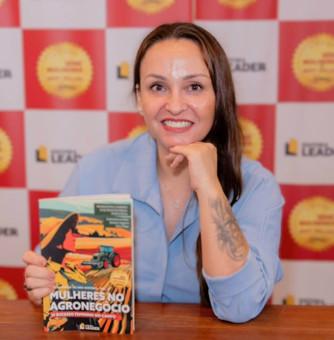 Cynthia M. Cominesi sorri segurando o livro Mulheres no Agronegócio, volume 2, diante do painel da Editora Leader.