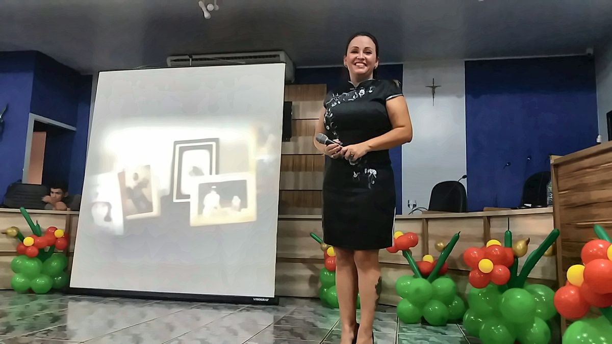 Cynthia M. Cominesi sorrindo no palco, com microfone na mão, ao lado do telão.
